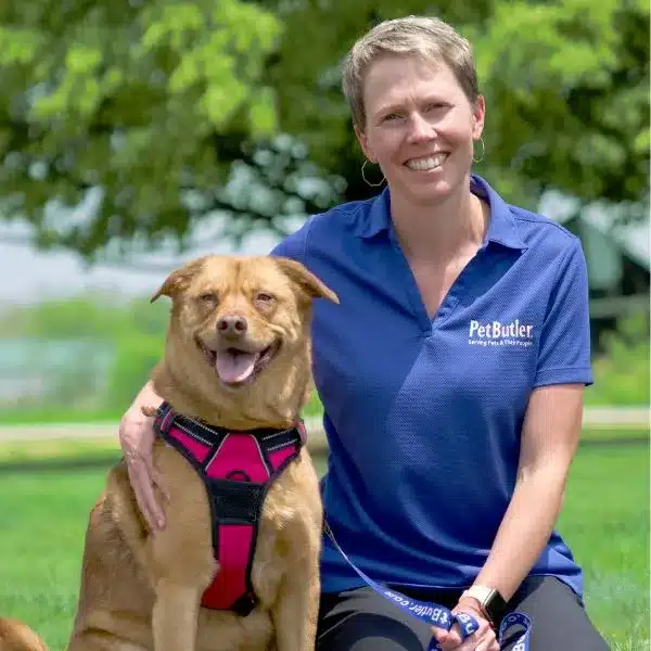 Pet Butler professional in a blue polo shirt kneeling in the grass next to a medium sized brown dog in a pink harness.