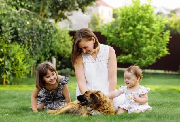 Woman sitting in the grass with a toddler girl and baby girl and petting a dog.