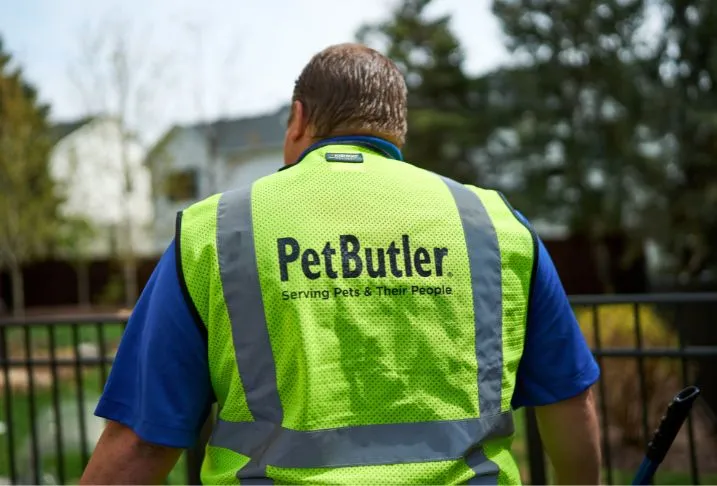 The back of a Pet Butler professional wearing a yellow Pet Butler logoed vest standing in a backyard.