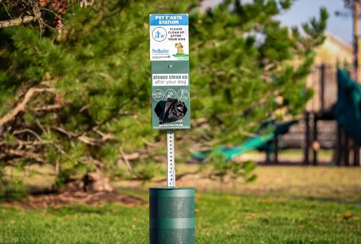 A Pet Butler pet waste station with black plastic bags in the dispenser and a garbage can at the base in a park.