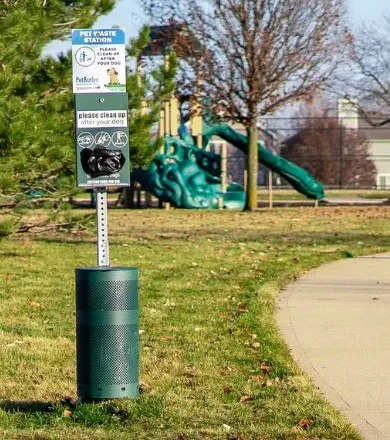 A Pet Butler pet waste station with a plastic bags dispenser and garbage can at the base next to a sidewalk in an open park area.