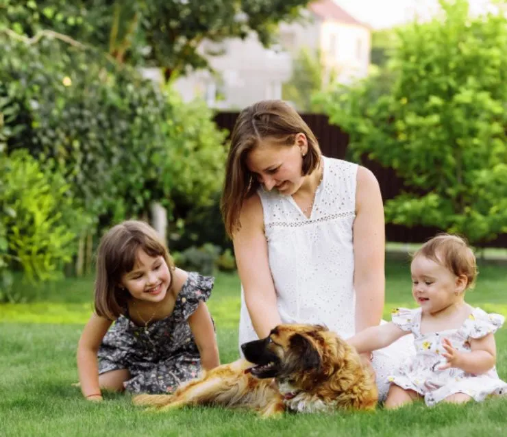 Woman sitting in the grass with a toddler girl and baby girl and petting a brown dog.