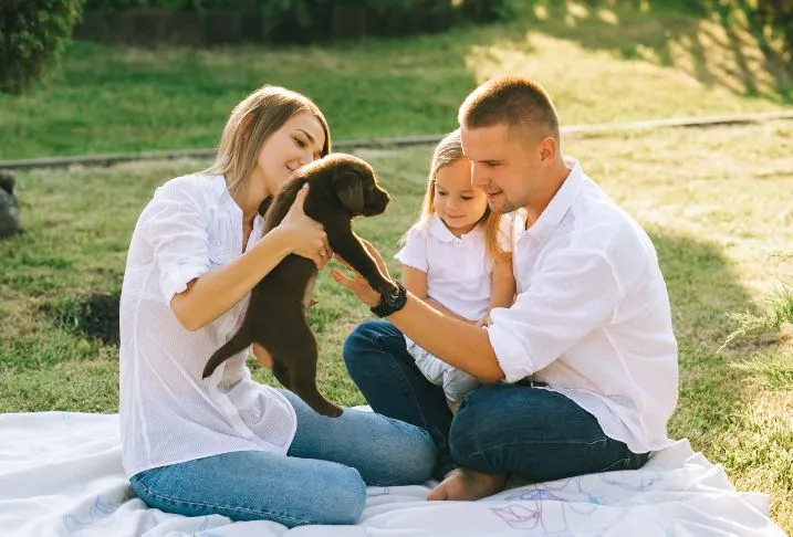 A man holding toddler girl and woman holding a black puppy sitting facing each other on a blanket on the grass.